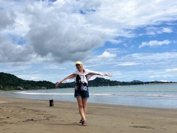 Innovations Academy founder Christine Kuglen stands with outstretched arms on a curving beach of sand with hills forested with trees in the background as a few shallow waves crash to shore.